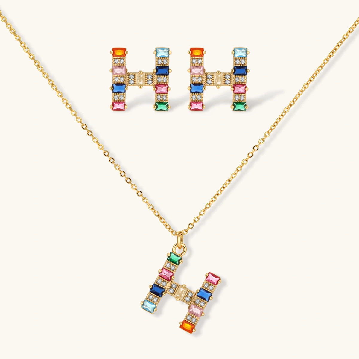 Gold-plated H initial pendant necklace and stud earrings set with multicolor baguette zirconia stones on cream background.