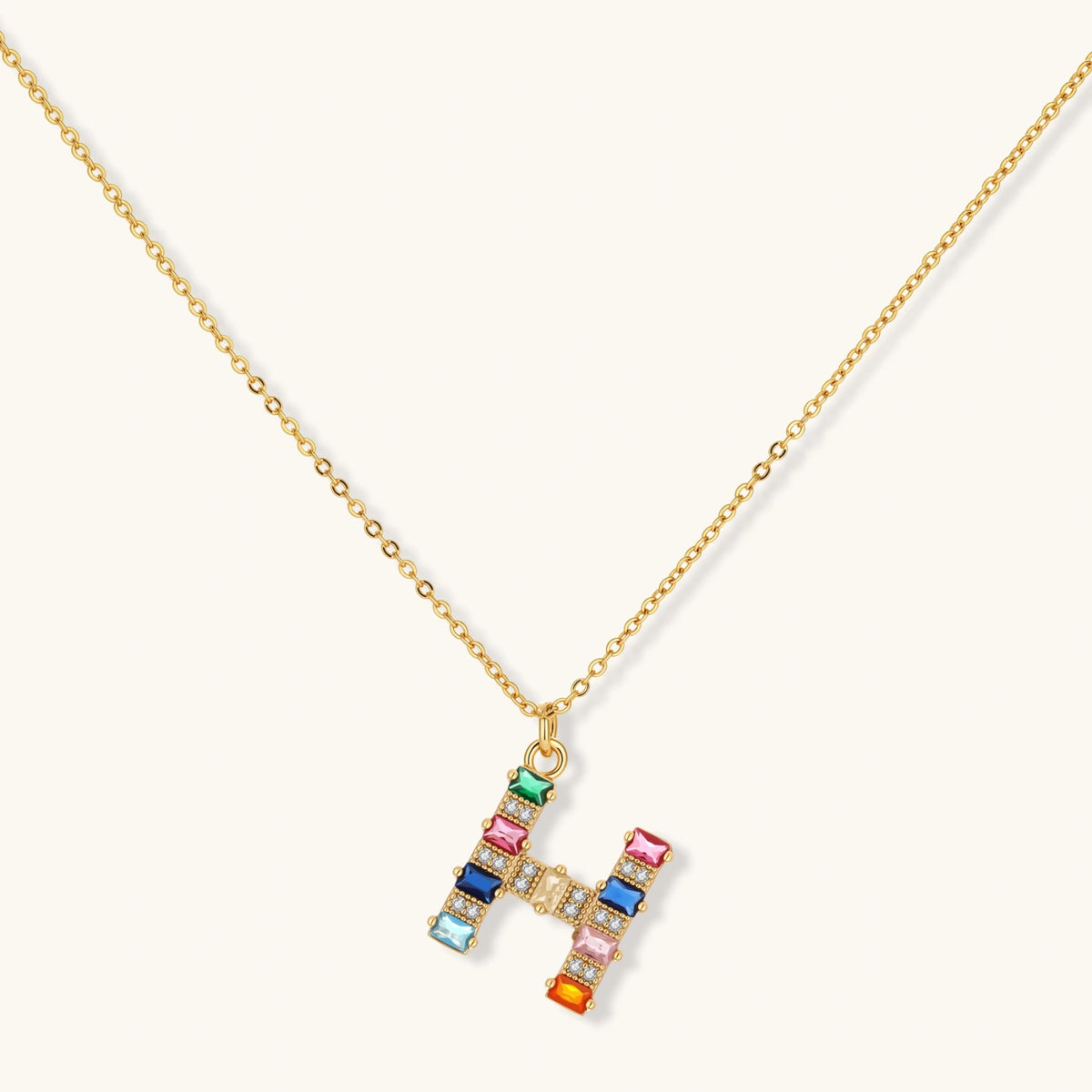 Gold-plated H initial pendant necklace and stud earrings set with multicolor baguette zirconia stones on cream background.